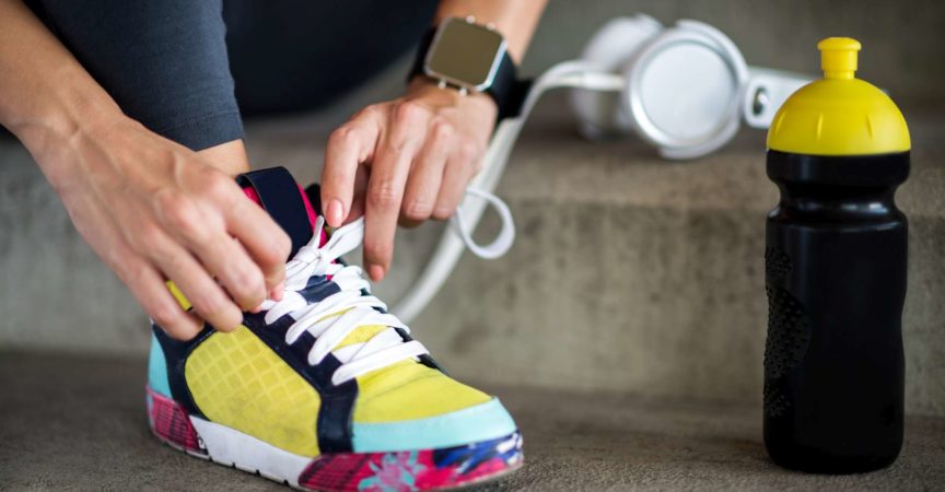 Weibliche Hände, die Schnürsenkel binden. Die Smartwatch befindet sich am linken Handgelenk und die Sportflasche befindet sich neben dem Sportschuh.