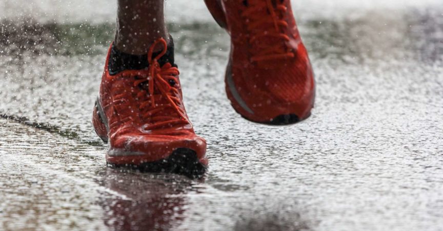 Nahaufnahme von Sportschuhen eines Athleten, der auf einem nassen Asphalt läuft. Spritzen Sie Wasser unter den Füßen hervor. Training im Regen.