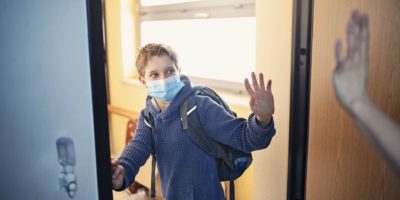 Kleiner Junge geht während der COVID-19-Pandemie zur Schule.
Mutter winkt ihm zum Abschied und er winkt zurück.
Nikon D850