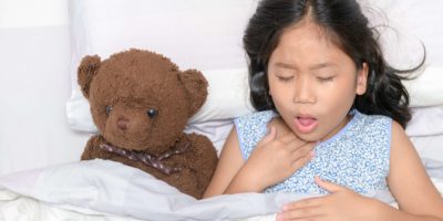 Kleines Mädchen hustet und Halsschmerzen, die auf Bett mit Spielzeugbär liegen, Gesundheitskonzept