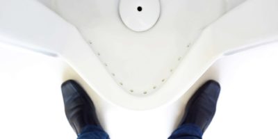 Draufsicht auf die Beine eines Mannes vor dem Urinal in der Herrentoilette