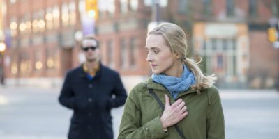 Verängstigte junge Frau mit blondem Haar, die über ihre Schulter auf einen Fremden in einem schwarzen Trenchcoat zurückblickt, der ihr in einer Großstadt folgt.