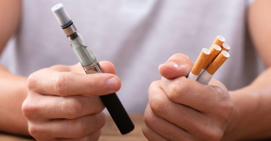 Man Holding Vape And Tobacco Cigarette Over Desk