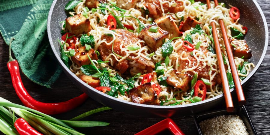 Süß-saurer Tofu, Hähnchenschenkel, chinesische Woknudeln, verwelkter Grünkohl, Sesam, roter Chili, Frühlingszwiebeln, serviert auf einer Pfanne mit Stäbchen auf dunklem Holzhintergrund, Draufsicht, Nahaufnahme