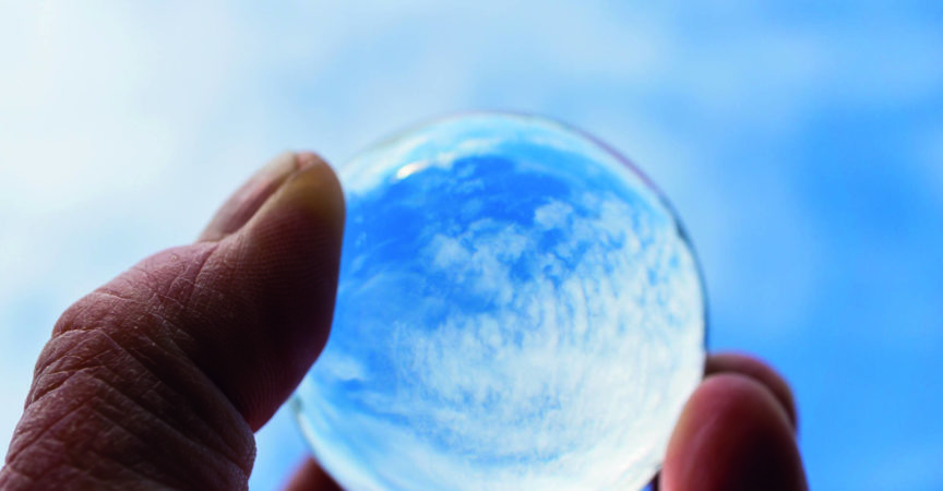 Die Hand eines Psoriasis-Patienten hält eine Glaskugel, in der sich ein blauer Himmel mit Wolken spiegelt. Ein Symbol der Hoffnung auf Genesung. Internationaler Psoriasis-Tag.