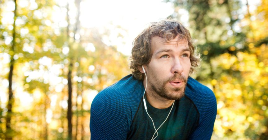 Junger hübscher Läufer mit Kopfhörern in seinen Ohren, Musik hörend, draußen in der sonnigen Herbstnatur, ruhend, ausatmend