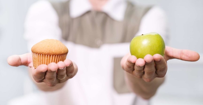 Doktor ein Zahnarzt ein Mann hält einen grünen Apfel und einen süßen Cupcake und zeigt auf der Waage eine Auswahl von nützlich gegen schädlich