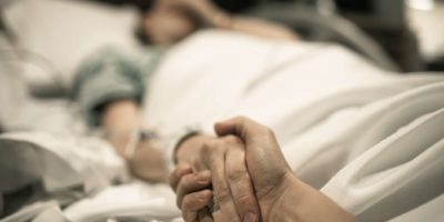Kranke Frau, die im Krankenhausbett mit der Hand liegt, die von der Liebe eins gehalten wird. Familienlinien und medizinisches Tragödienkonzept.