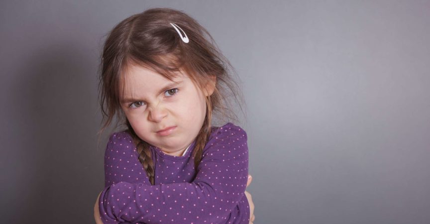 Ein kleines süßes Mädchen schmollt. Sie trägt ein lila Hemd vor einem grauen Hintergrund.