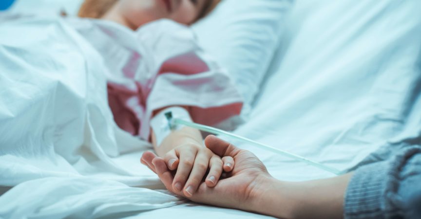 Das kleine Kind, das im Krankenhausbett schläft, erholt sich, ihre Hand fällt in die Mutter und sie hält sie tröstend. Konzentrieren Sie sich auf die Hände. Emotionaler Familienmoment.