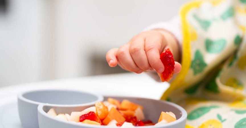 Makro Nahaufnahme der Babyhand mit einem Stück Obst, das im Kinderstuhl sitzt Kind isst gesundes Essen