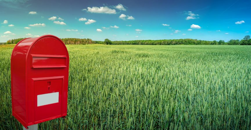 Großer roter moderner Briefkasten mit weißem leerem Notizraum für Adresse steht im Freien vor schönem Landschaftshintergrund mit grünem Weizenfeld und blauem Himmel.