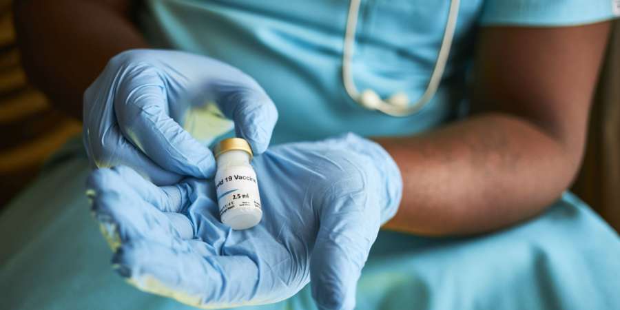 Ein schwarzafrikanischer Arzt, der mit chirurgischem Peeling und Handschuhen sitzt und eine Durchstechflasche mit Covid-19-Impfstoff in seinen Händen hält