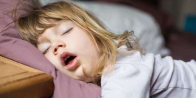 lustiger Gesichtsausdruck mit offenem Mund eines blonden, kaukasischen dreijährigen Kindes, das auf einem Kingsize-Bett schläft