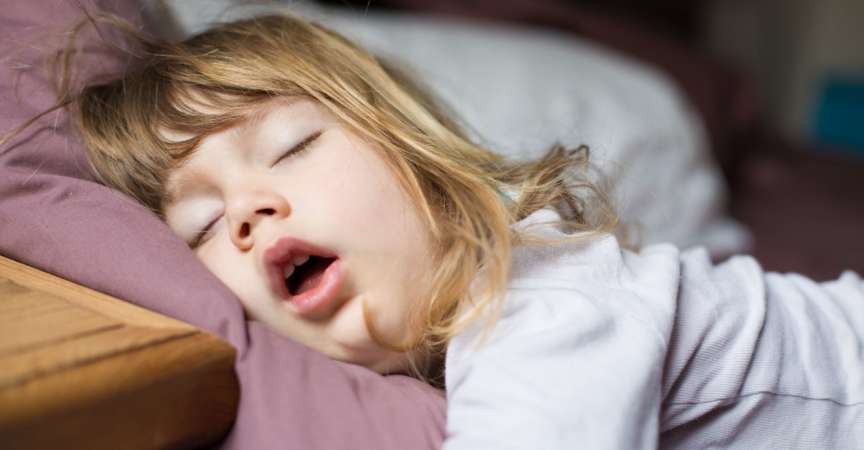 lustiger Gesichtsausdruck mit offenem Mund eines blonden, kaukasischen dreijährigen Kindes, das auf einem Kingsize-Bett schläft