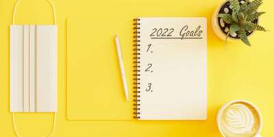 2022 Neujahrsziele Konzept. Draufsicht auf Schutzmaske, Topfpflanze und Kaffeetasse auf gelbem Hintergrund