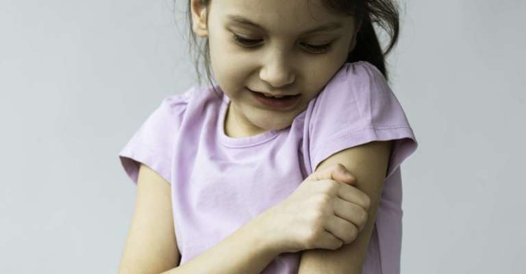 Kleines Mädchen kratzt sich vor grauem Hintergrund am Arm.