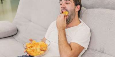 ein mann sitzt auf einer couch mit einer schuessel chips auf dem schoss und isst chips