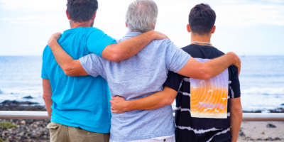 Familiengruppe, drei Männer, Vater, erwachsener Sohn und Teenager-Enkel, die sich umarmen und den Horizont über Wasser betrachten