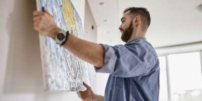 Mann mit Bart, der in seinem Wohnzimmer ein Gemälde an die Wand hängt