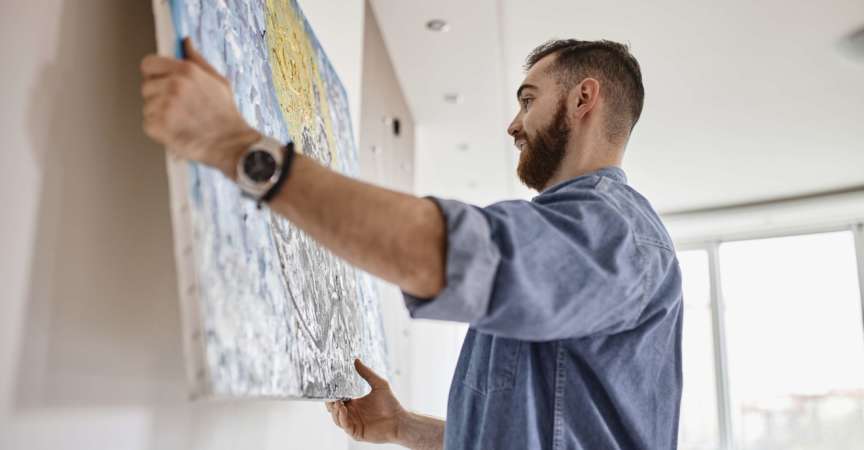 Mann mit Bart, der in seinem Wohnzimmer ein Gemälde an die Wand hängt
