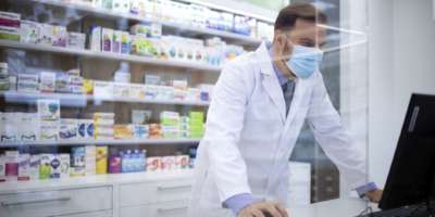 Apotheker mit Gesichtsschutzmaske und weißem Mantel, der während der Corona-Virus-Pandemie Vitamine im Apothekenladen verkauft.