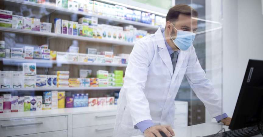 Apotheker mit Gesichtsschutzmaske und weißem Mantel, der während der Corona-Virus-Pandemie Vitamine im Apothekenladen verkauft.