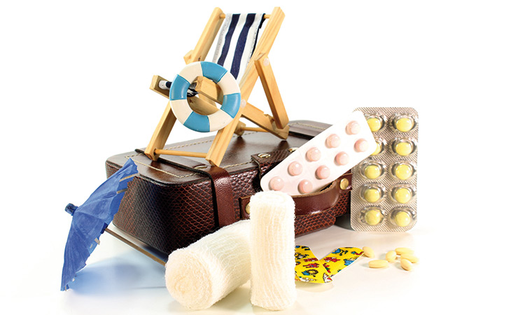 Ein Reiseapotheke-Beutel mit diversen Medikamenten, Pflaster sowie einem Mini-Liegestuhl aus Holz vor weissem Hintergrund