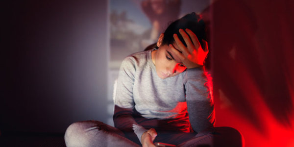 Ein Teenager Mädchen sitzt im Schneidersitz am Boden im Dunkeln vor einer, den Kopf in die eine Hand gestützt, in der anderen hält sie ihr Handy. Ein Projektor projiziert Bilder eines Social Media Feeds auf sie und die Rückwand.