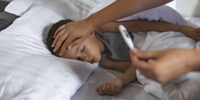Foto eines kranken männlichen Kindes mit hohem Fieber, das im Bett liegt, während die Mutter die Temperatur misst.