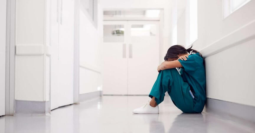 Gestresste und überarbeitete Ärztin, die Scrubs trägt, sitzt auf dem Boden im Krankenhauskorridor