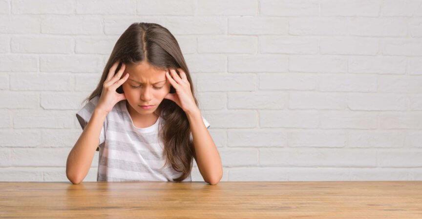 Junges hispanisches Kind, das zu Hause auf dem Tisch sitzt, mit der Hand auf dem Kopf, weil es Stress im Kopf hat. Migräne leiden.