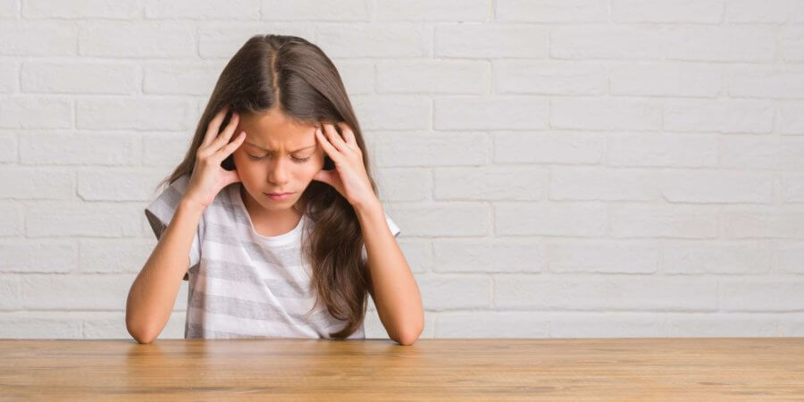 Junges hispanisches Kind, das zu Hause auf dem Tisch sitzt, mit der Hand auf dem Kopf, weil es Stress im Kopf hat. Migräne leiden.