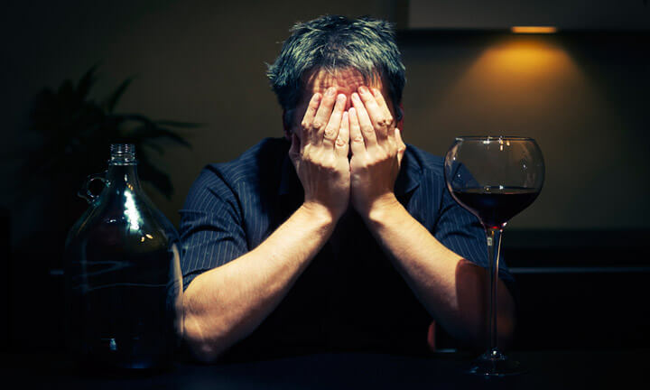 Betrunkener sitzt an einem Tisch in einem dunklen Raum mit einer fast leeren Flasche Rotwein und versteckt das Gesicht in den Händen.