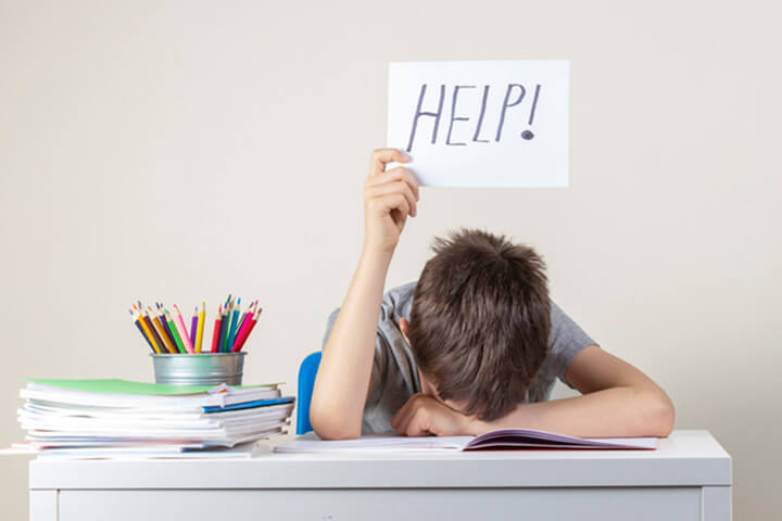 Trauriger, müder, frustrierter Junge, der mit vielen Büchern am Tisch sitzt und Papier mit dem Wort „Hilfe“ in der Hand hält.
