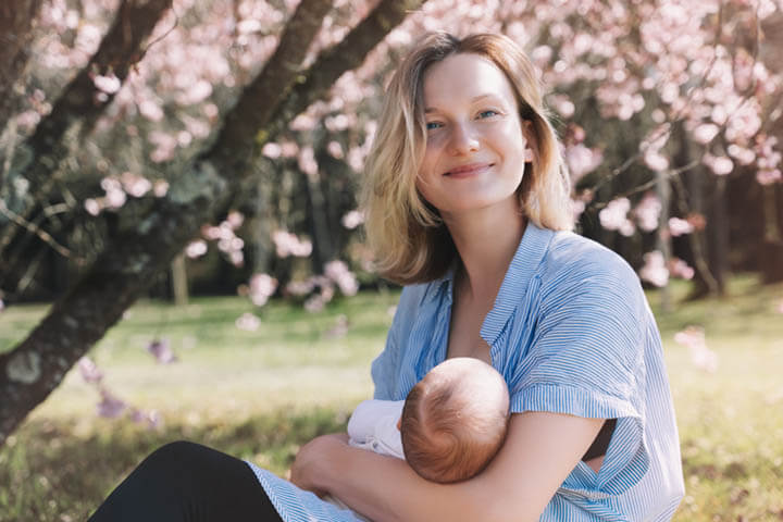 Junge Frau stillt ihr neugeborenes Baby im Freien unter einem Baum und lacht in die Kamera.