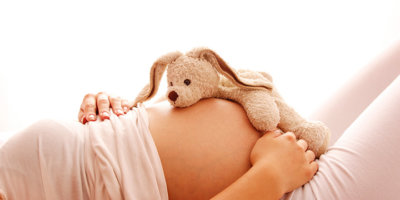 Teddybär liegt auf dem Bauch einer schwangeren Frau vor weissem Hintergrund.
