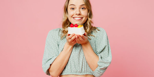 Junge fröhliche Frau vor pinkem Hintergrund hält mit beiden Händen ein Stück Kuchen in die Höhe.