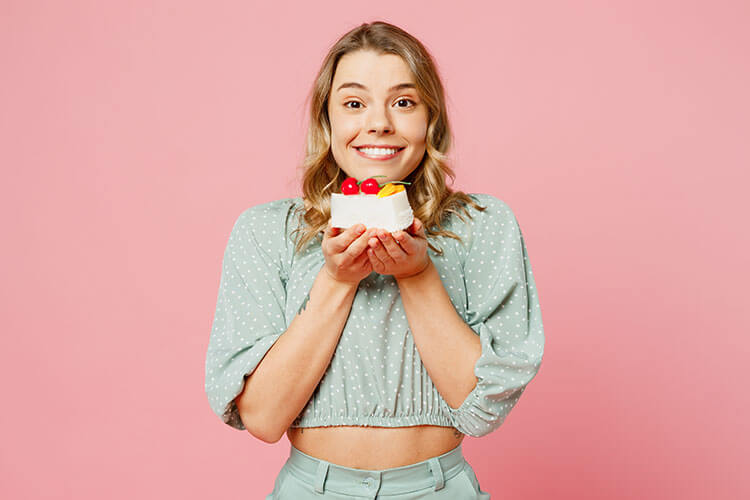 Junge fröhliche Frau vor pinkem Hintergrund hält mit beiden Händen ein Stück Kuchen in die Höhe.