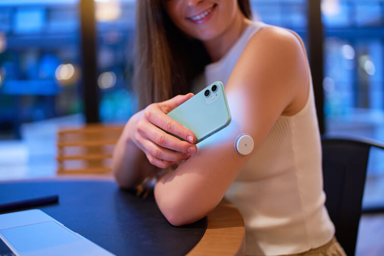 Junge Frau an einem Tisch misst Blutzucker mit Smartphone über Sensor am Oberarm