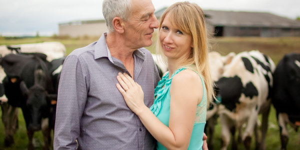 Älterer Mann umarmt seine jüngere blonde Frau auf einer Kuhwiese.