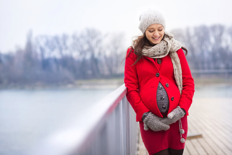 Zufriedene schwangere Frau steht im Winter gut eingepackt auf einer Brücke.